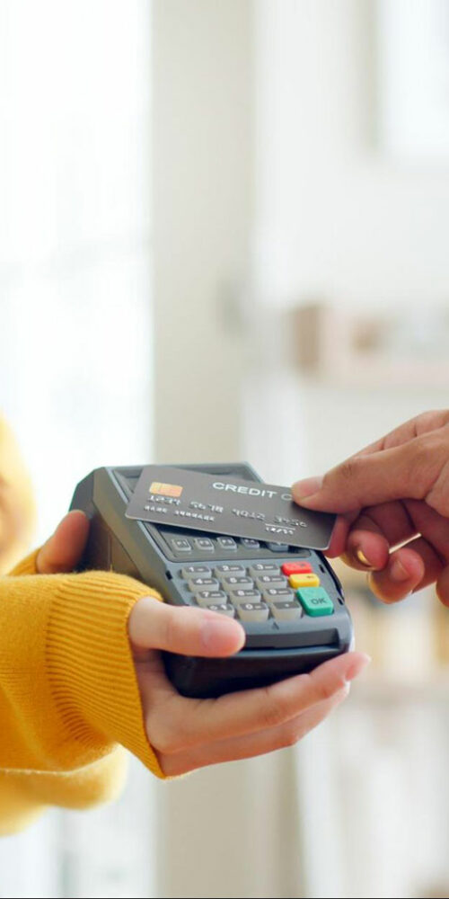 An image of a person in a yellow sweater holding a credit card payment terminal, and another person's hand is inserting a credit card into the slot of the terminal. The background is blurred, and it appears to be an indoor setting.