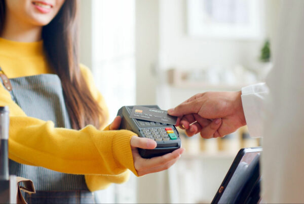 An image of a person in a yellow sweater holding a credit card payment terminal, and another person's hand is inserting a credit card into the slot of the terminal. The background is blurred, and it appears to be an indoor setting.