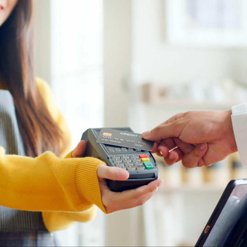 An image of a person in a yellow sweater holding a credit card payment terminal, and another person's hand is inserting a credit card into the slot of the terminal. The background is blurred, and it appears to be an indoor setting.