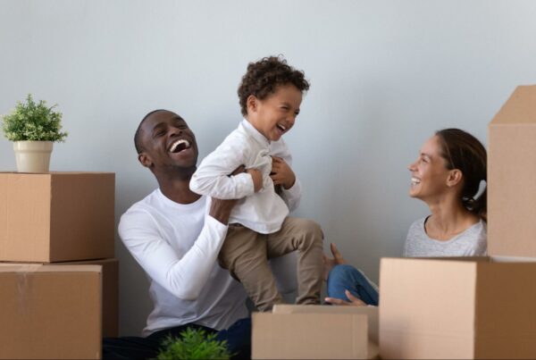 A joyful family moment with a father lifting a laughing toddler in the air while the mother sits nearby, all surrounded by moving boxes, suggesting they are in the process of moving into a new home.