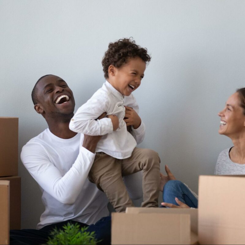 A joyful family moment with a father lifting a laughing toddler in the air while the mother sits nearby, all surrounded by moving boxes, suggesting they are in the process of moving into a new home.