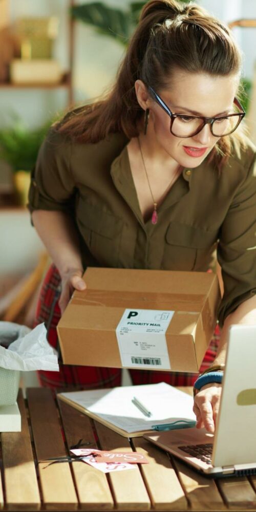 A woman in a green shirt and glasses packing a box amidst various packing materials, with a laptop open in front of her on a wooden table, and clothing items hanging on a rack in the background, indicating a small business workspace.