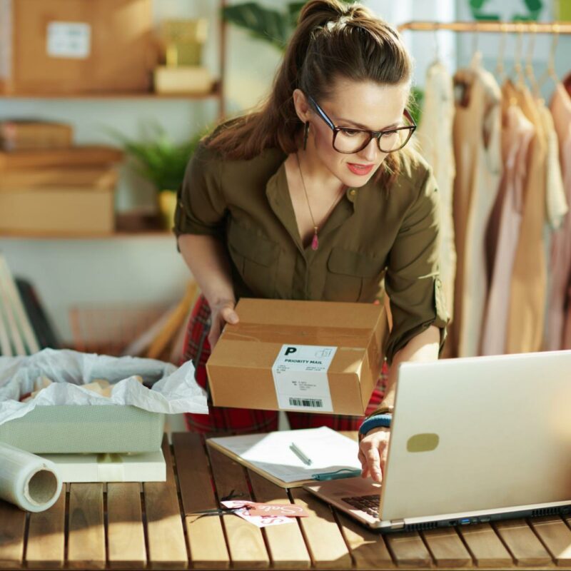 A woman in a green shirt and glasses packing a box amidst various packing materials, with a laptop open in front of her on a wooden table, and clothing items hanging on a rack in the background, indicating a small business workspace.
