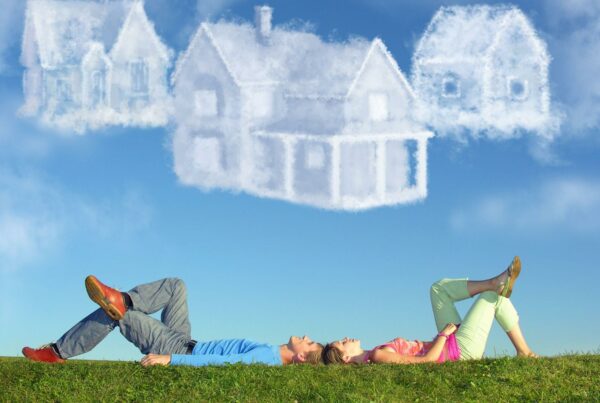 A whimsical image of a man and woman lying back-to-back on a grassy hill, gazing upwards at clouds shaped like various styles of houses. The sky is clear and blue, creating a stark contrast with the white, fluffy cloud houses above them.