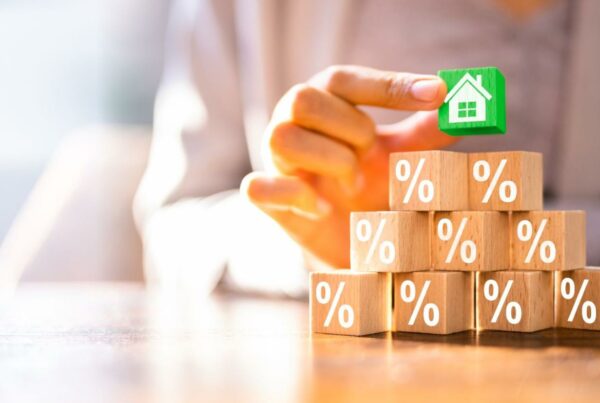 A person's hand placing a small, green house figurine on top of stacked wooden blocks, each marked with a percentage symbol, symbolizing mortgage rates or financing options.